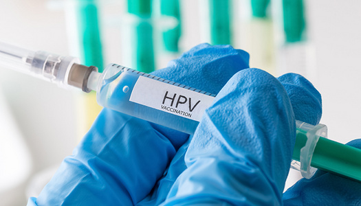 Hand mit blauem Latexhandschuh hält Impfspritze mit Aufschrift "HPV"